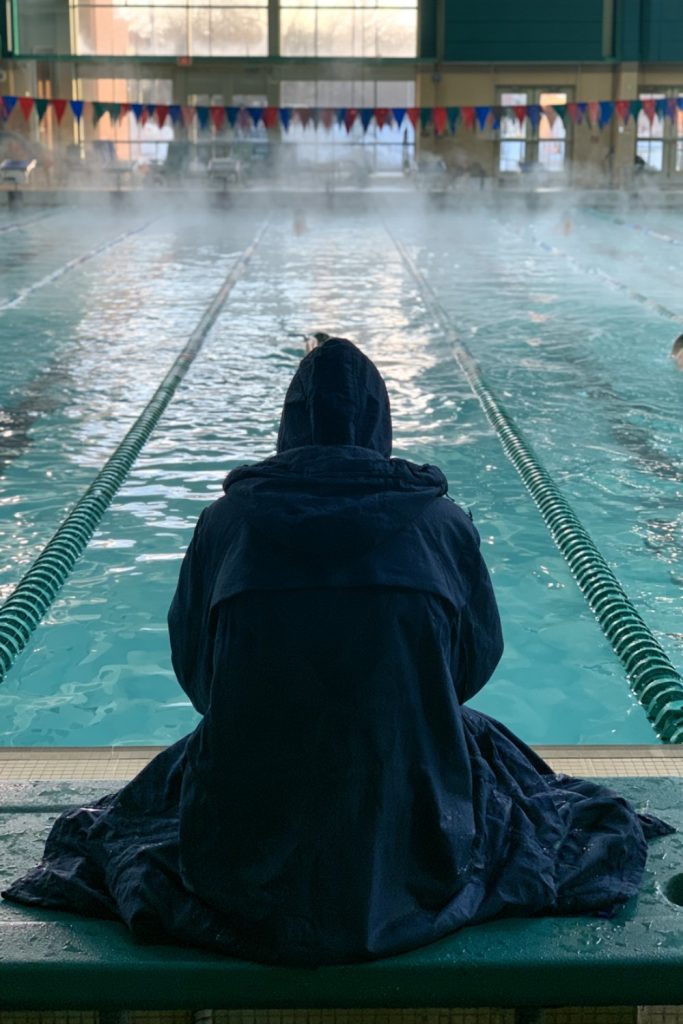 Swimmer in a long navy parka seen from behind on a pool deck bench facing the lanes