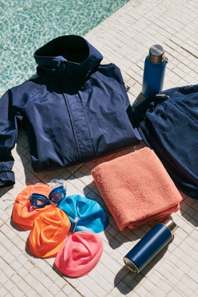 Navy parka, bright swim caps, goggles and a towel laid out beside a pool