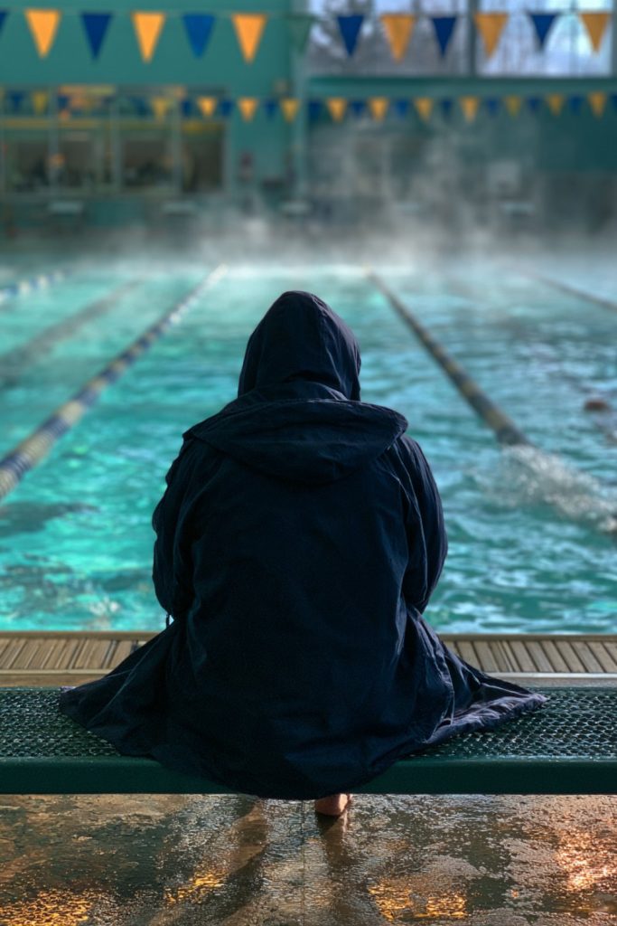 Swimmer wrapped in a parka sitting on a bench at an indoor pool with backstroke flags overhead