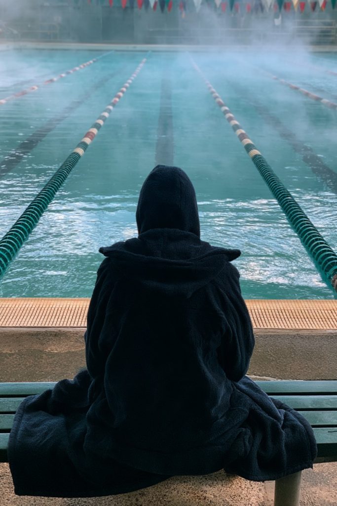 Swimmer seen from behind in a hooded parka on the pool deck with steam over the water