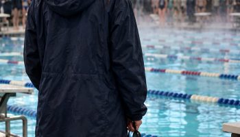Swimmer seen from behind on an indoor pool deck in a long navy parka holding goggles