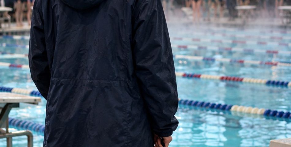Swimmer seen from behind on an indoor pool deck in a long navy parka holding goggles