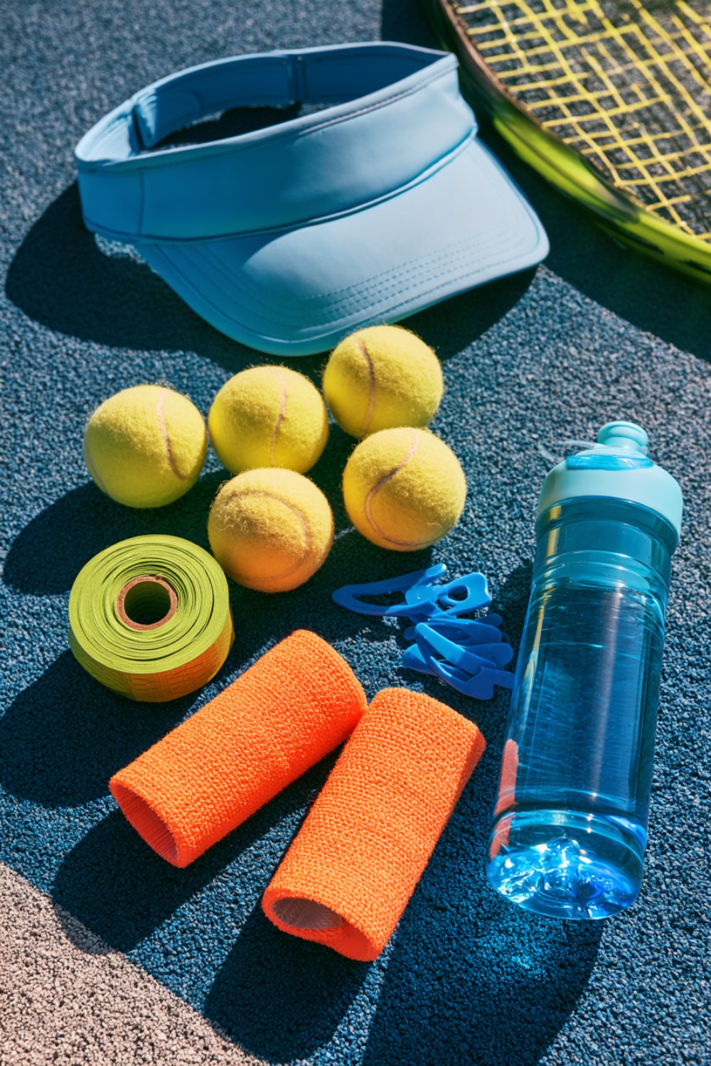 Tennis gifts on a blue court: a visor, yellow balls, orange wristbands, overgrip and a water bottle