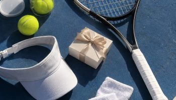 Flat lay on a blue hard court with a racquet, an open can of yellow balls, wristbands, a white visor and a small gift box