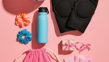 Flat lay of volleyball gift ideas on a pink background: a blue and white volleyball, black knee pads, scrunchies, a water bottle, striped socks and a mesh drawstring bag
