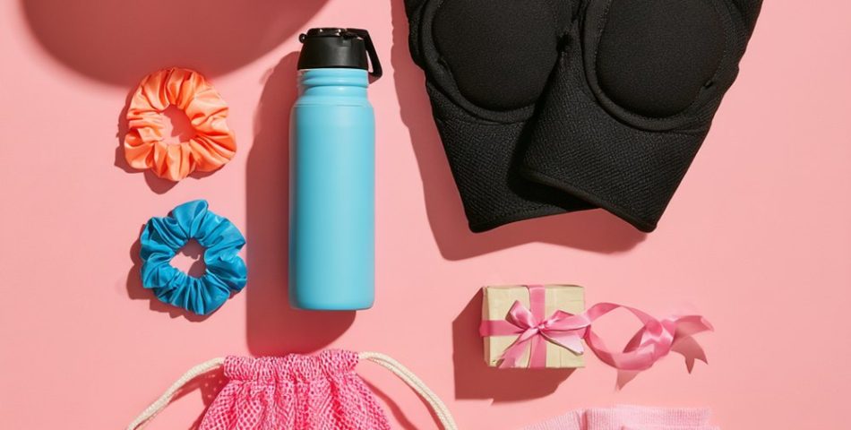 Flat lay of volleyball gift ideas on a pink background: a blue and white volleyball, black knee pads, scrunchies, a water bottle, striped socks and a mesh drawstring bag