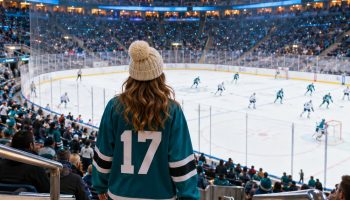 Hockey game outfit idea: woman seen from behind in an oversized teal hockey jersey, black leggings, white sneakers and cream beanie walking down arena stairs