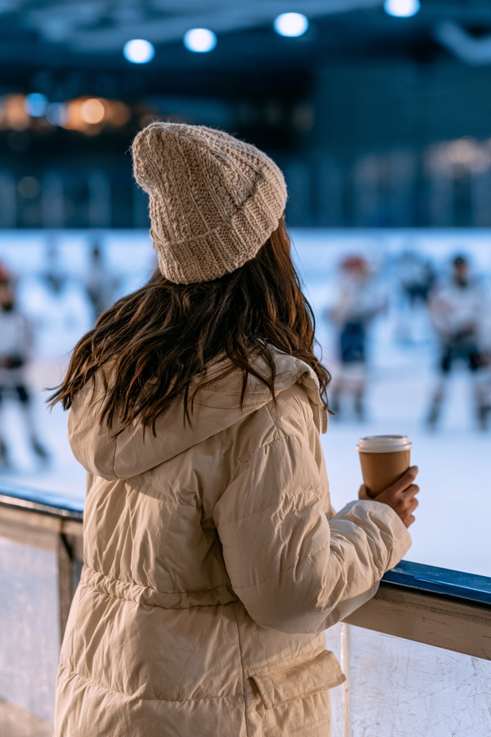 Cozy rink outfit from behind: long cream puffer coat, knit beanie and a hot coffee at the boards