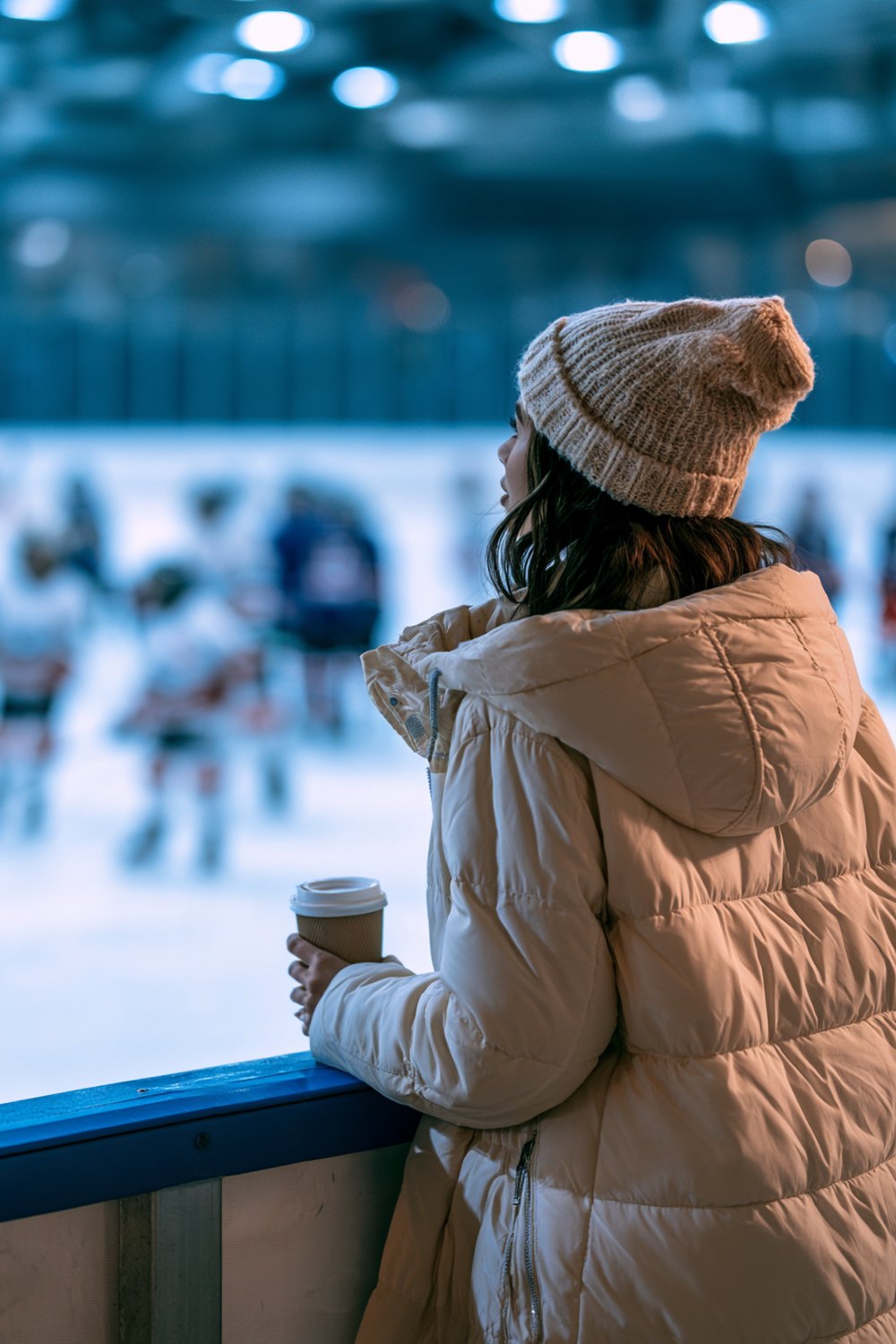 Hockey mom seen from behind at the rink glass in a cream puffer coat and knit beanie holding a coffee