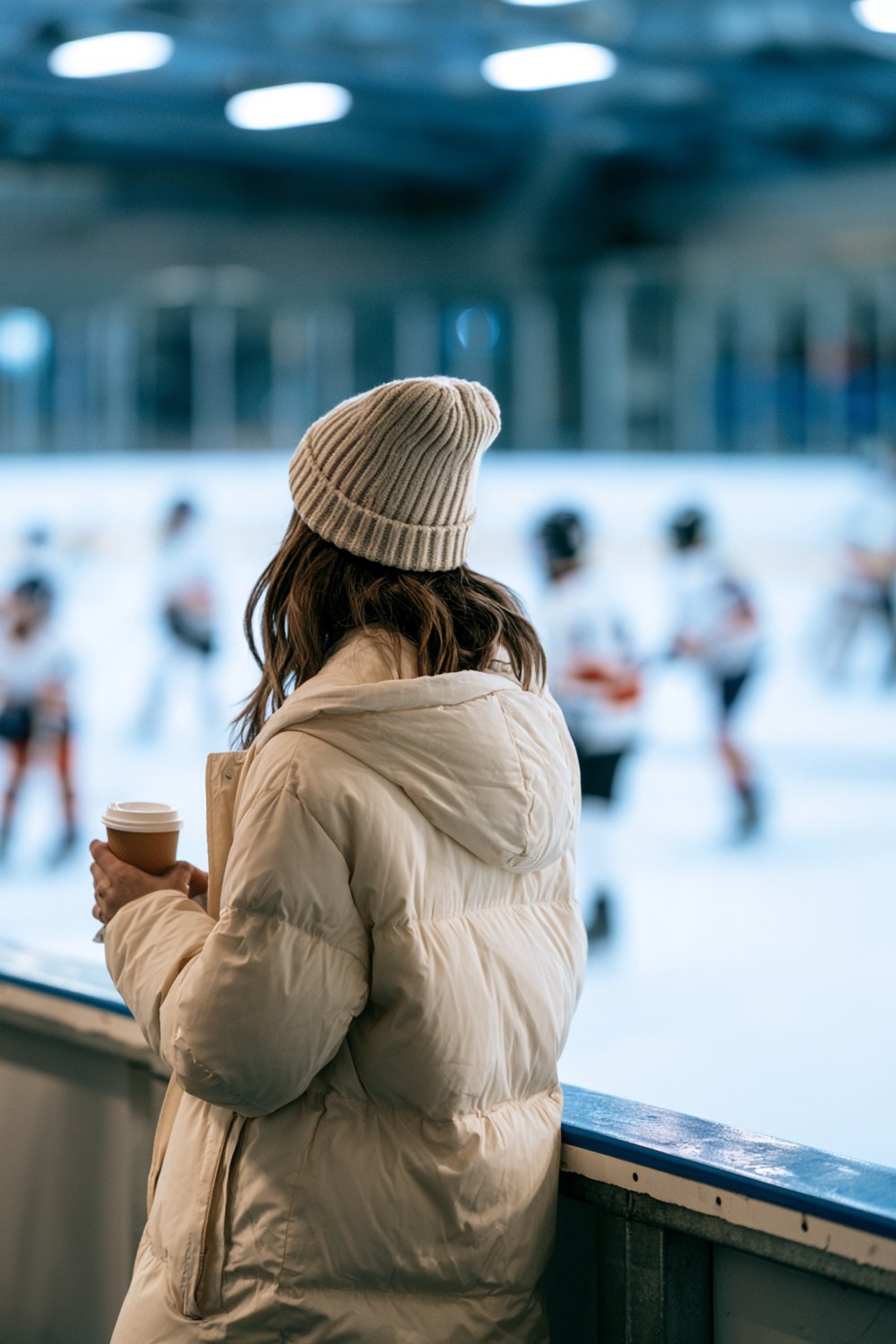 Woman from behind in a cream puffer and beanie watching youth hockey at an indoor rink