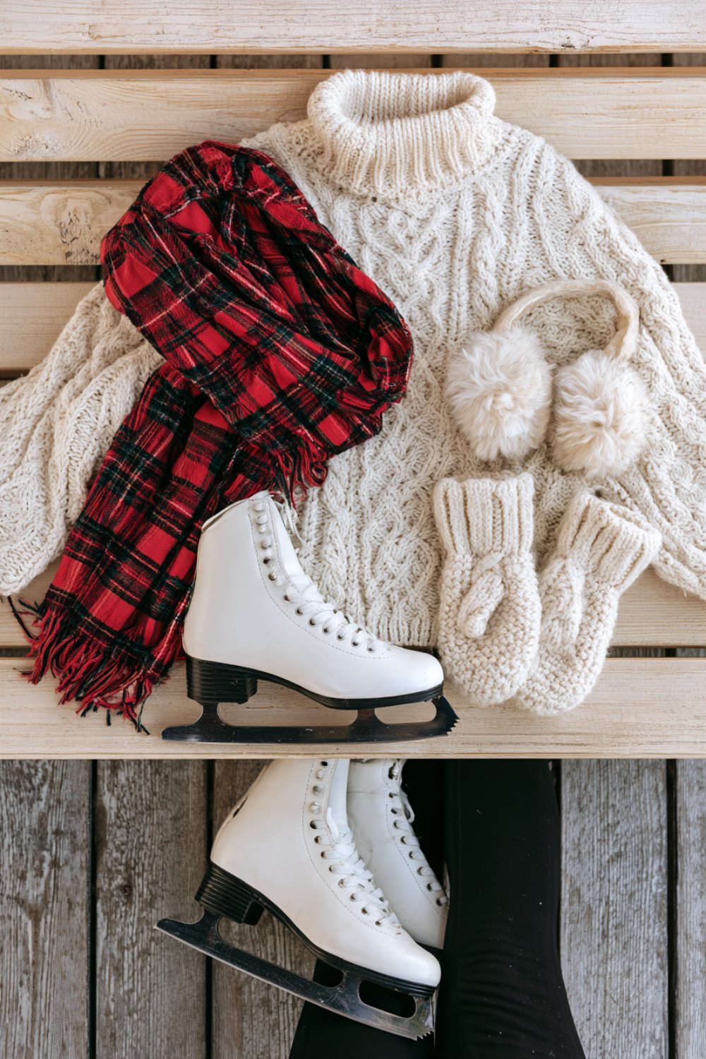 Cream knit sweater, plaid scarf, white skates, fluffy earmuffs and mittens laid on a pale wooden bench