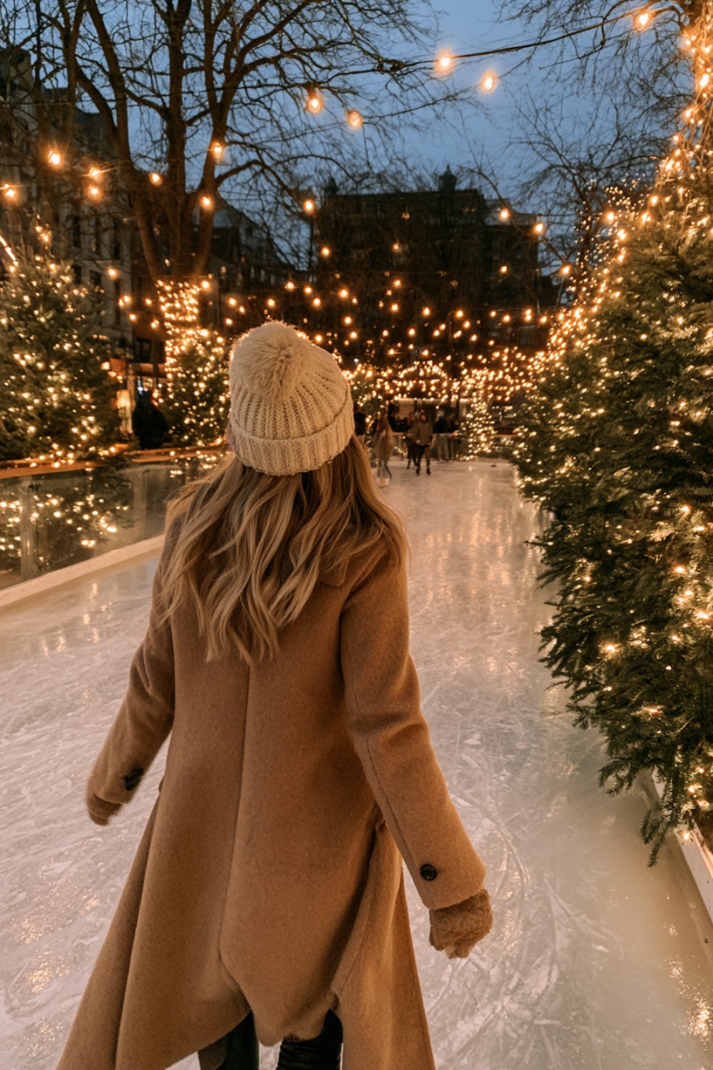 Person in a long camel coat and cream beanie skating away on an outdoor rink with string lights