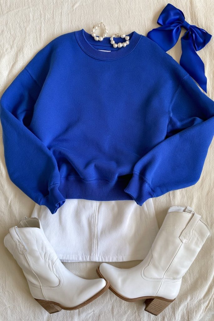 Flat lay of a royal blue crewneck sweatshirt with a blue hair bow and pearl earrings on cream linen