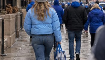 Fan in a light blue puffer and grey beanie carrying a clear bag on a wet downtown street, seen from behind