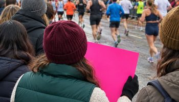 Spectators seen from behind along an autumn city marathon course, one in an emerald puffer vest holding a bright pink sign