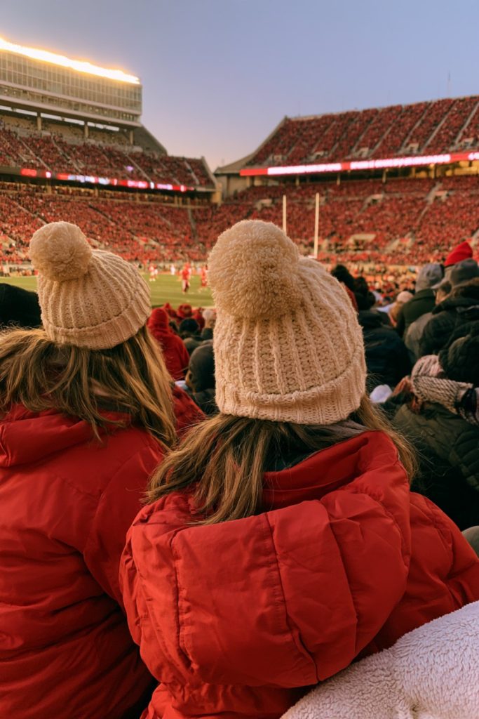 Fans in cream beanies seen from behind in a stadium full of red under evening lights