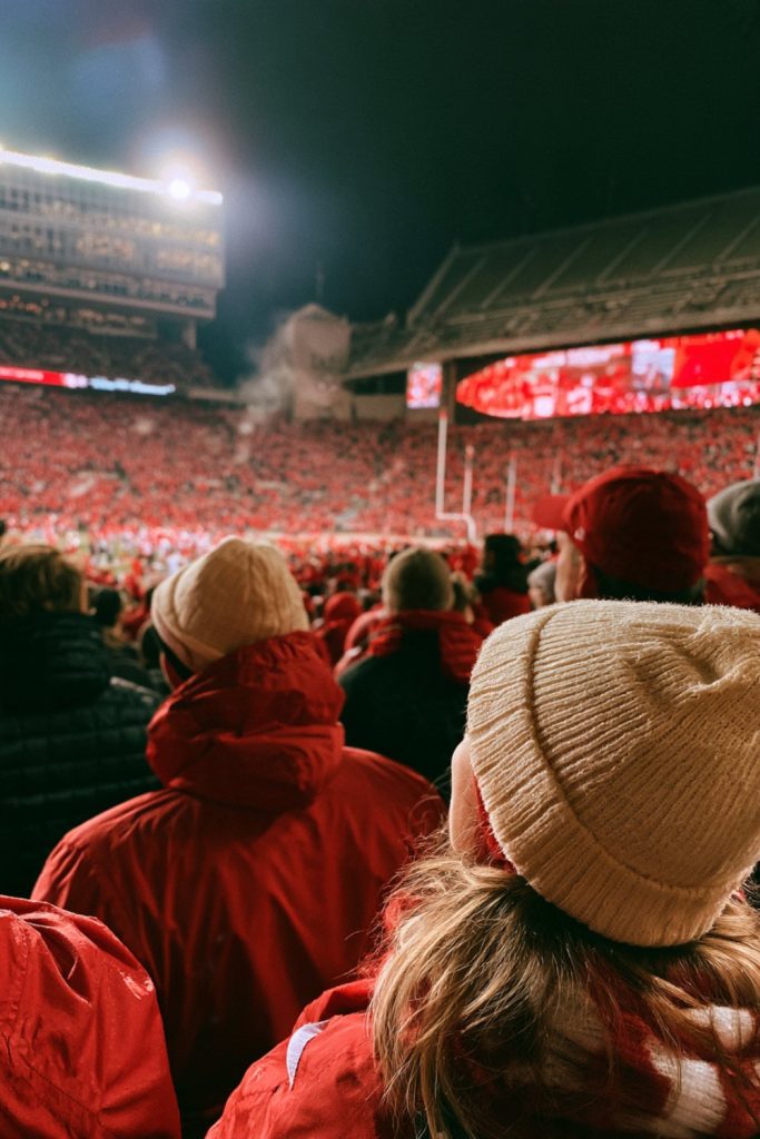 Crowd in red jackets and cream hats seen from behind at a night college football game