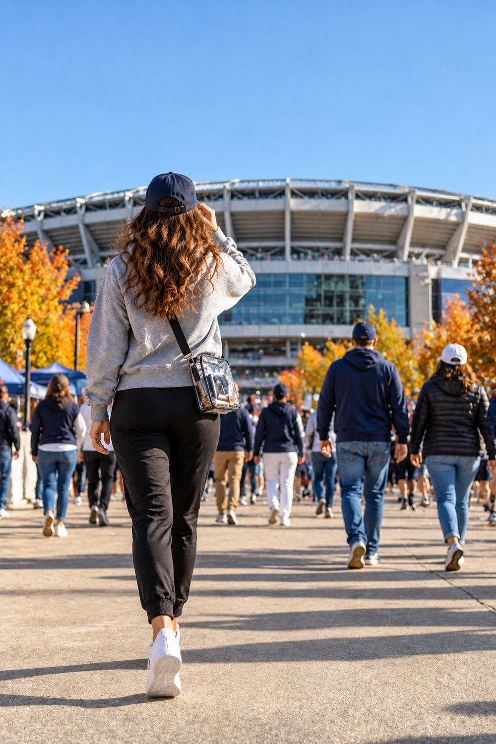 Woman walking to a football stadium in a grey hoodie, black joggers, white sneakers, cap and clear crossbody bag