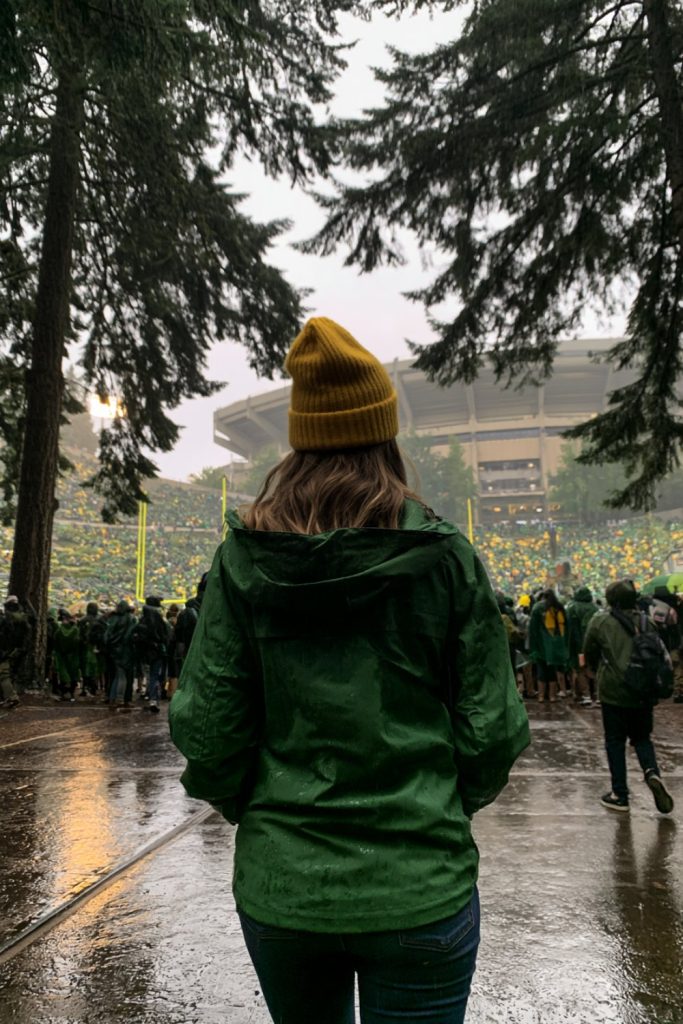 Woman in a green rain jacket and yellow beanie walking through tall evergreens toward a stadium in the rain