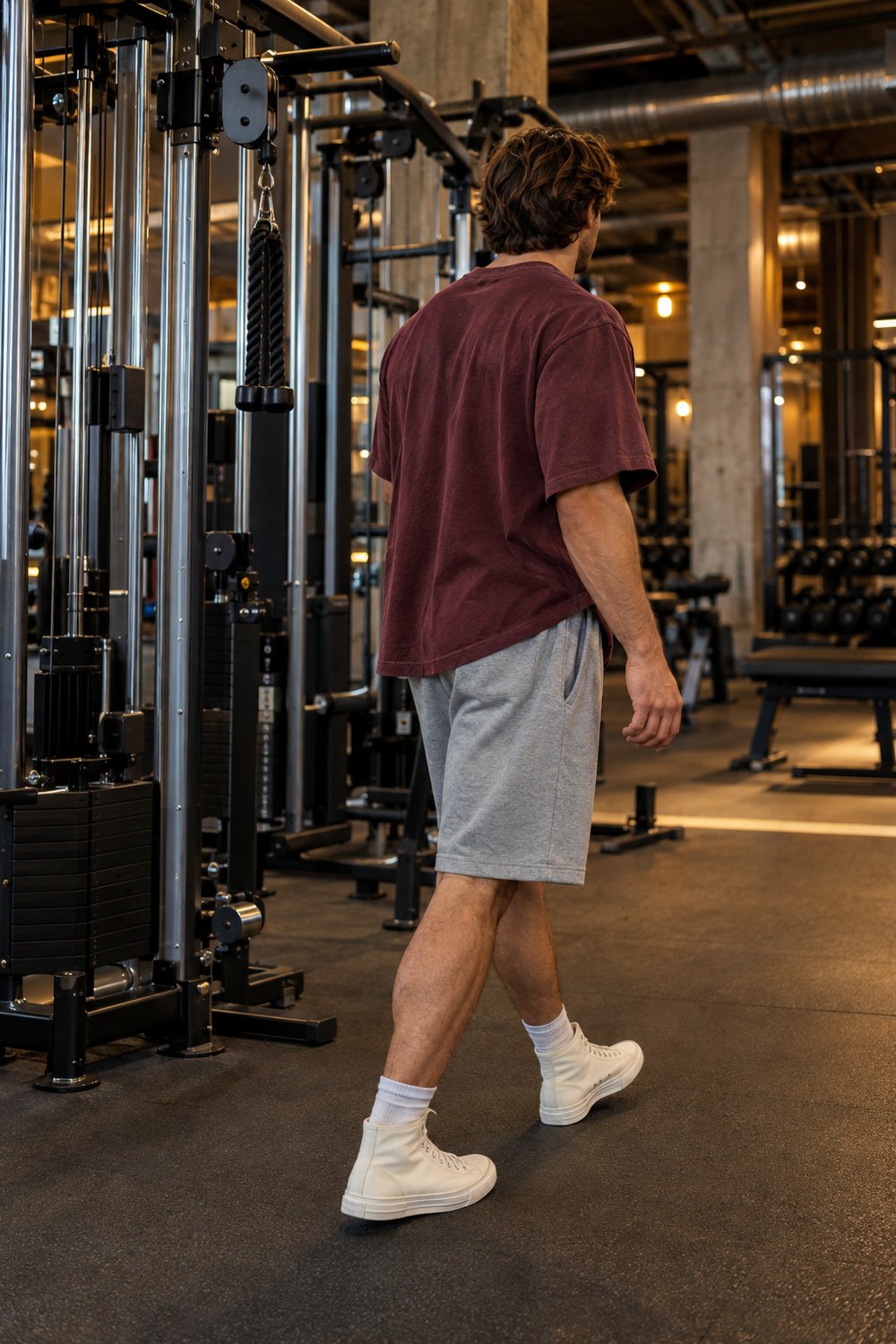 Man in an oversized burgundy t-shirt, grey sweat shorts and off-white canvas high tops walking past cable machines