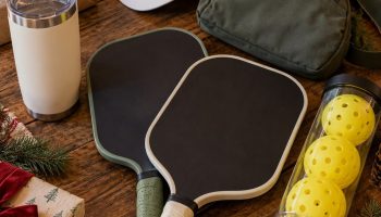 Christmas flat lay of pickleball gifts: two paddles, a tube of balls, a sling bag, a visor, a tumbler and wrapped boxes