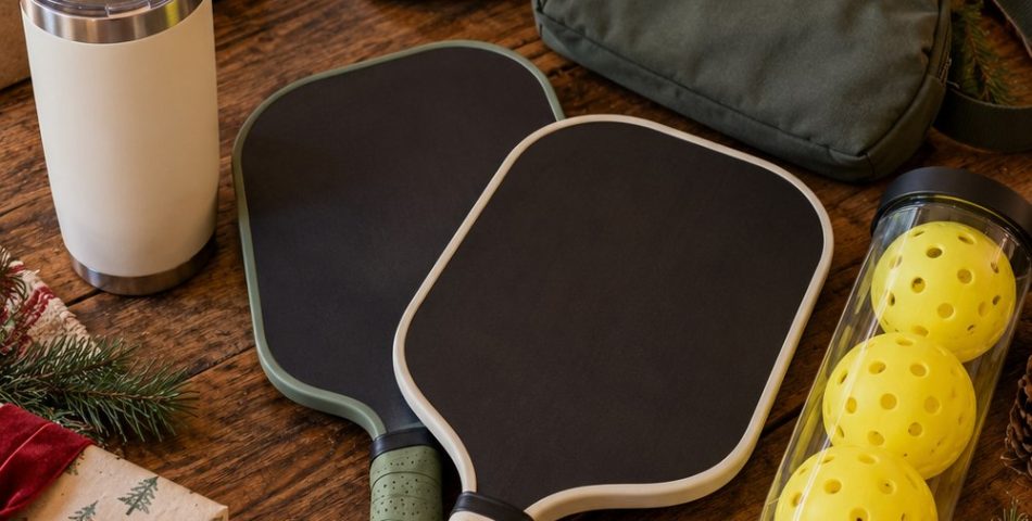 Christmas flat lay of pickleball gifts: two paddles, a tube of balls, a sling bag, a visor, a tumbler and wrapped boxes