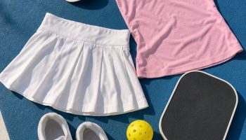 Flat lay of a pickleball outfit on a blue court: white pleated skort, pink tank, white court sneakers, visor, paddle and ball