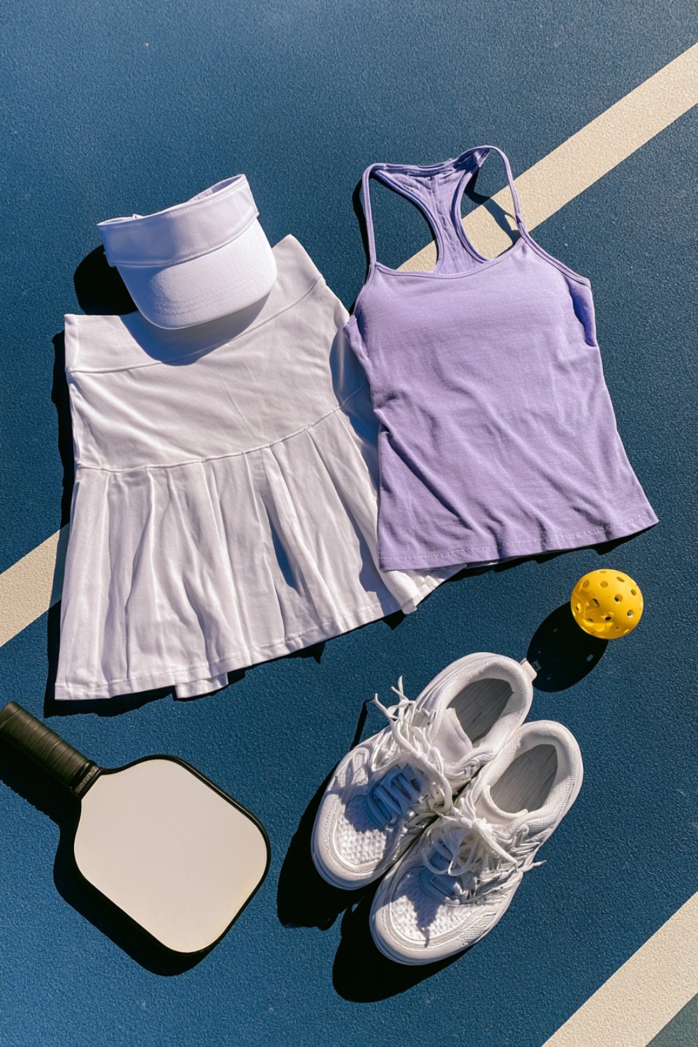 White pleated skort and lavender tank with white court sneakers, a visor and paddle laid on court lines
