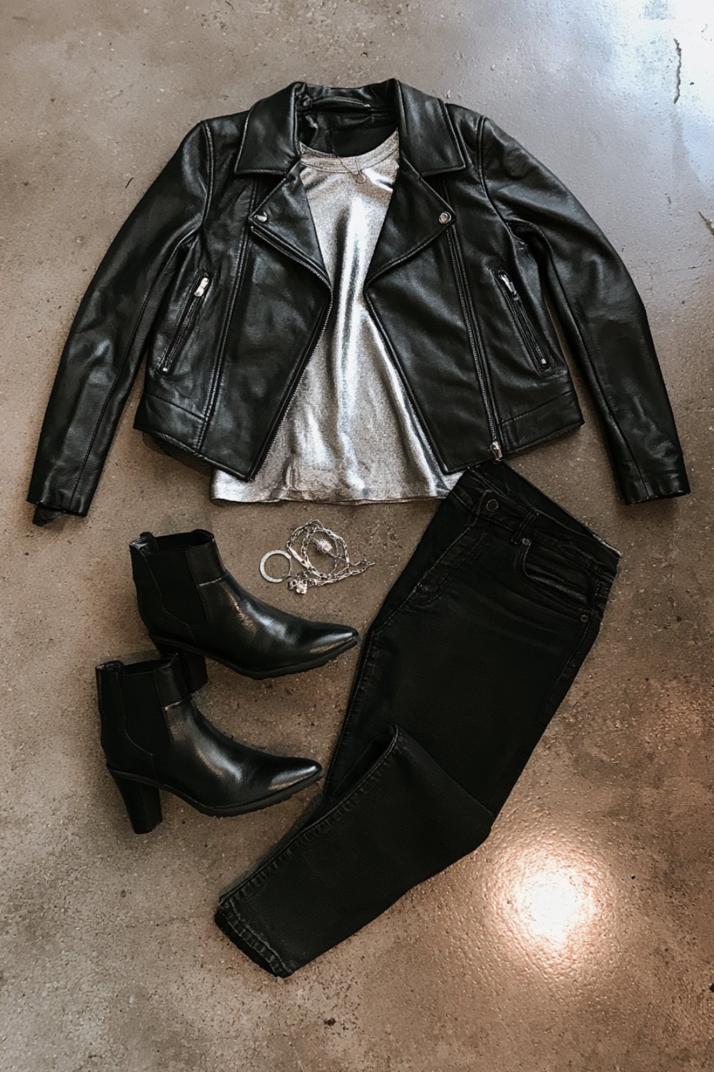 Flat lay of a black leather moto jacket, silver top, black jeans, black ankle boots and silver jewelry on concrete