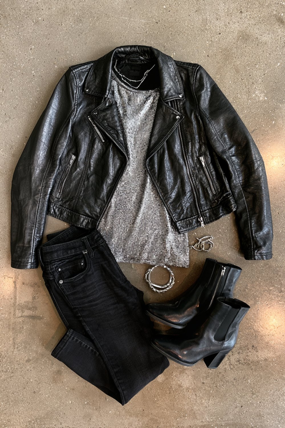 Black leather jacket and silver metallic top with black jeans and boots laid out on a concrete floor