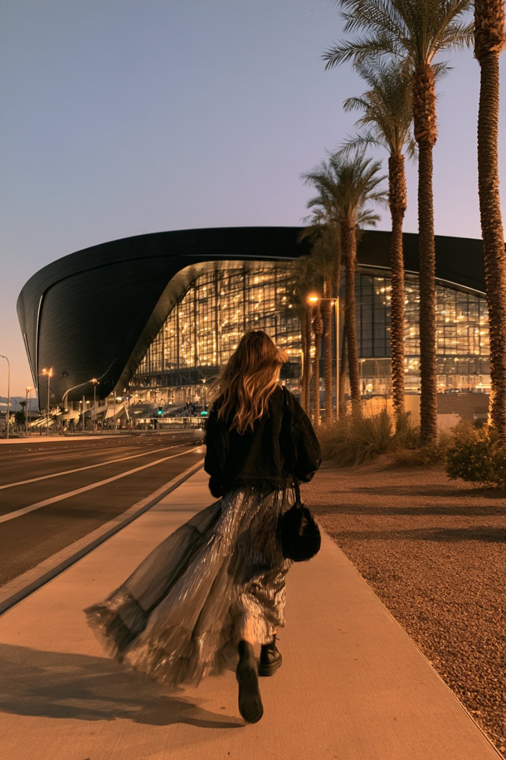 Woman seen from behind in a black top and metallic skirt walking toward a dark modern stadium at dusk