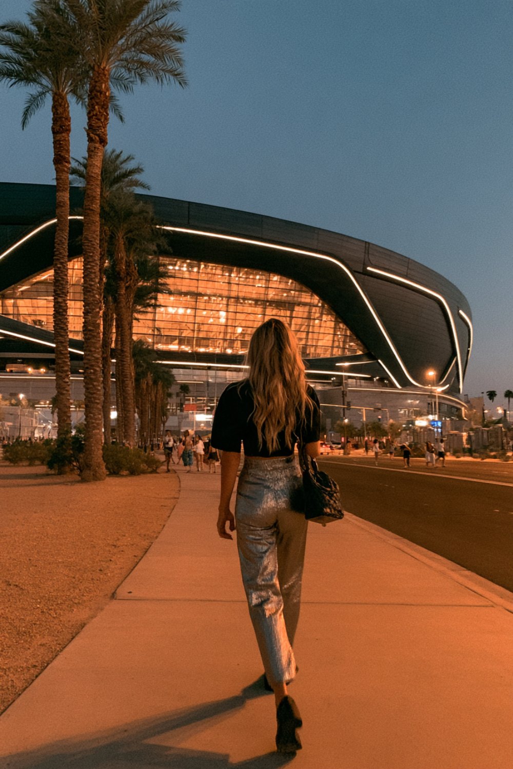 Woman from behind in a black top and silver pants on a sidewalk outside a sleek black stadium at sunset