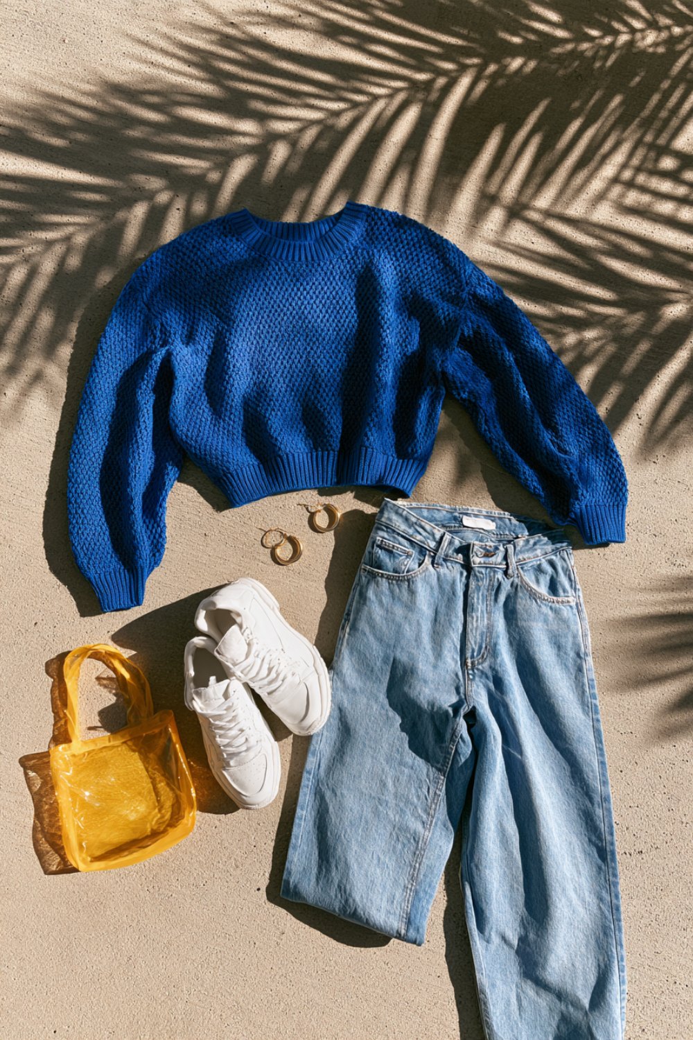 Flat lay of a cropped royal blue sweater, light wash jeans, white sneakers and a yellow bag on a sunlit patio