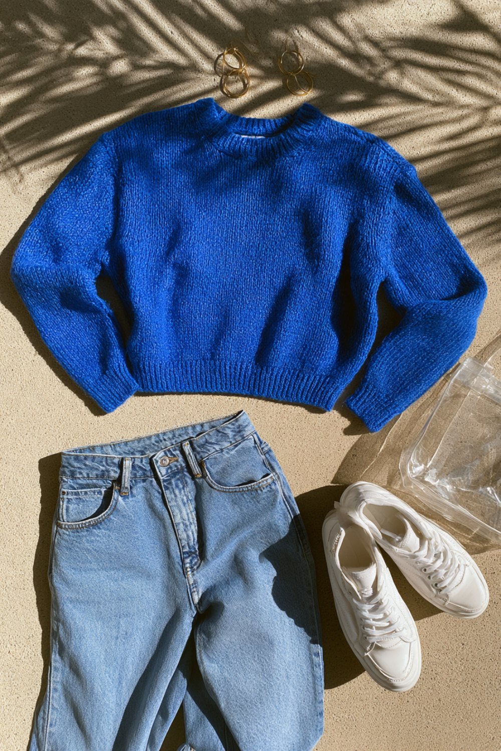 Royal blue knit sweater, straight jeans and white sneakers flat lay with a small clear bag