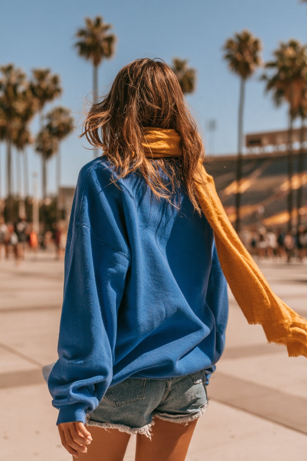 Woman seen from behind in a royal blue sweatshirt and golden yellow scarf walking across a palm-lined stadium plaza