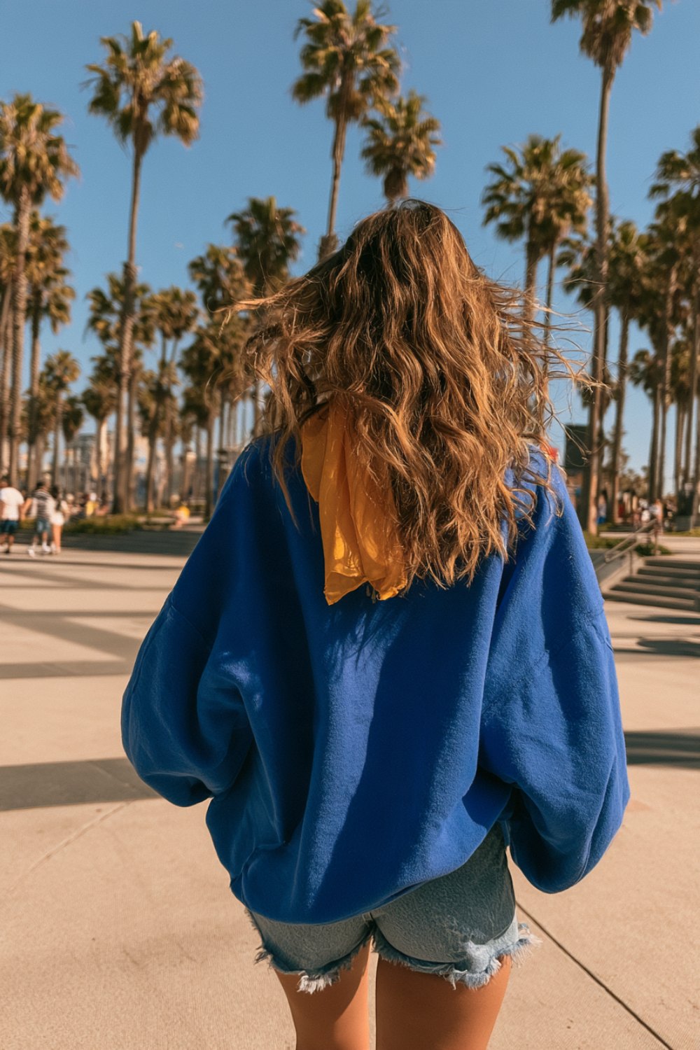 Woman from behind in an oversized royal blue sweatshirt and denim shorts on a sunny Southern California plaza