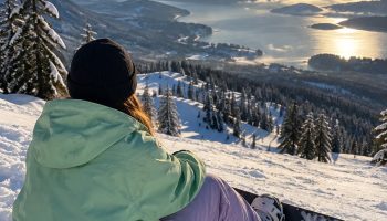 Snowboarder seen from behind sitting on fresh snow in a mint green jacket and lavender pants with a mountain view