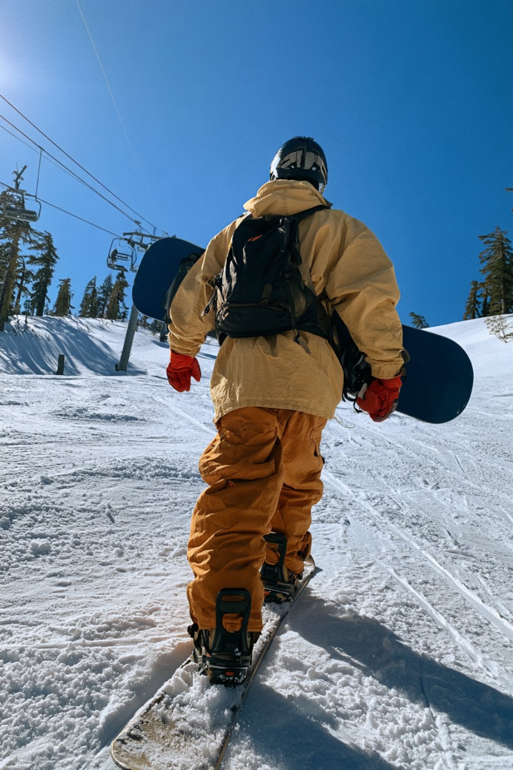 Snowboarder in mustard snow pants and a black jacket walking toward a chairlift