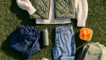 Flat lay on green turf of an olive quilted vest, cream knit sweater, light jeans, white sneakers, rain shell and orange slices