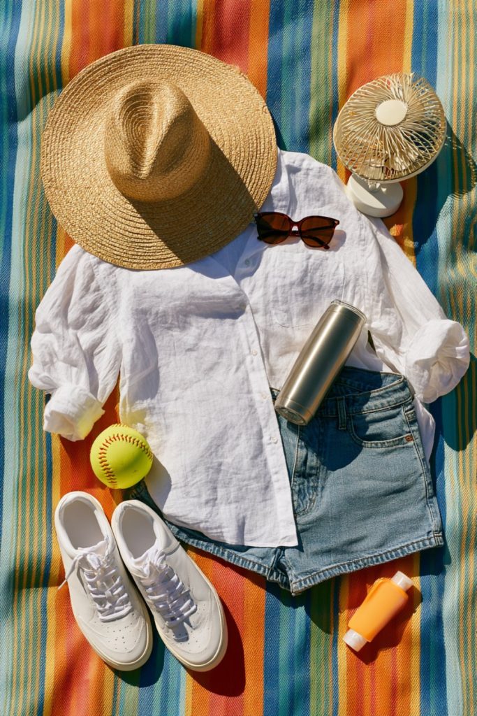 Straw hat, linen shirt, denim shorts, tumbler and softball laid out on a bright striped picnic blanket