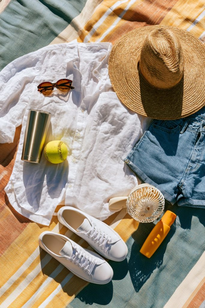 Hot weather softball mom outfit flat lay with a sun hat, sunglasses, sneakers and an insulated tumbler