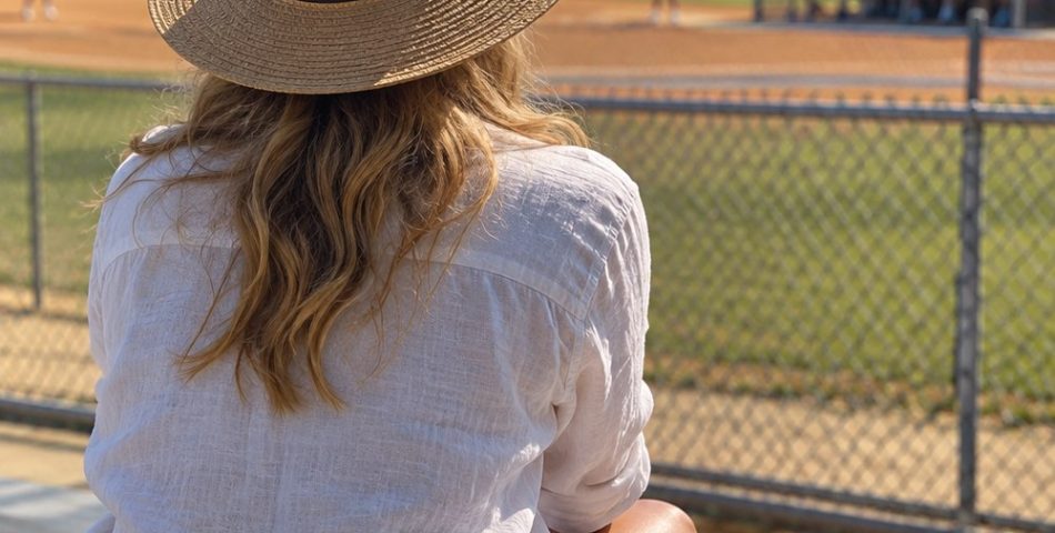 Softball mom seen from behind on aluminum bleachers in a white linen shirt, denim shorts and straw hat, tumbler beside her
