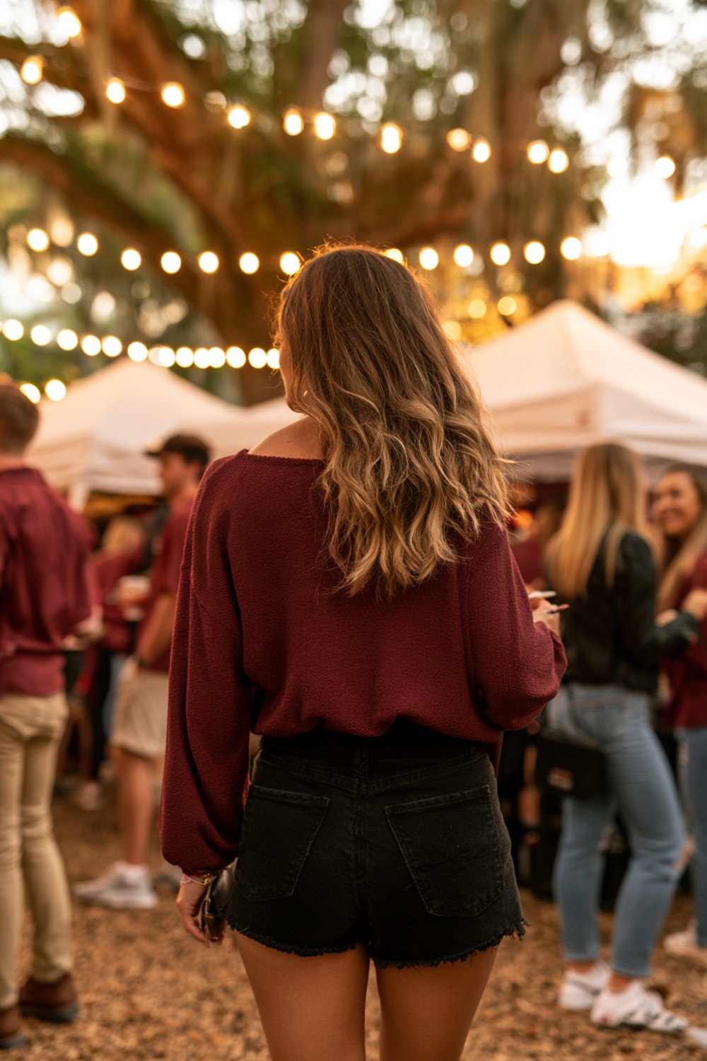 Woman in a garnet top seen from behind at a college tailgate under oak trees and string lights