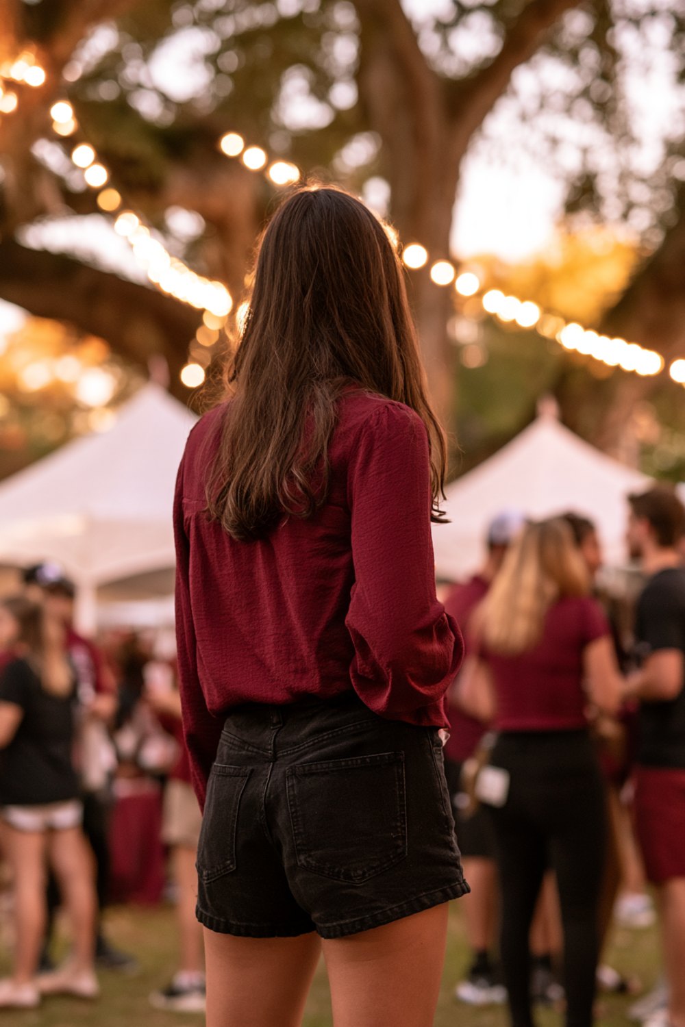 Woman in a garnet top seen from behind at a college tailgate under oak trees and string lights, wider view