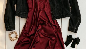 Flat lay on a white porch of a garnet satin dress, black denim jacket, black boots, gold hoops and a clear stadium bag