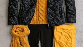 Flat lay of a black quilted puffer jacket, mustard gold cable knit sweater, black leggings and boots with a plain gold towel on concrete