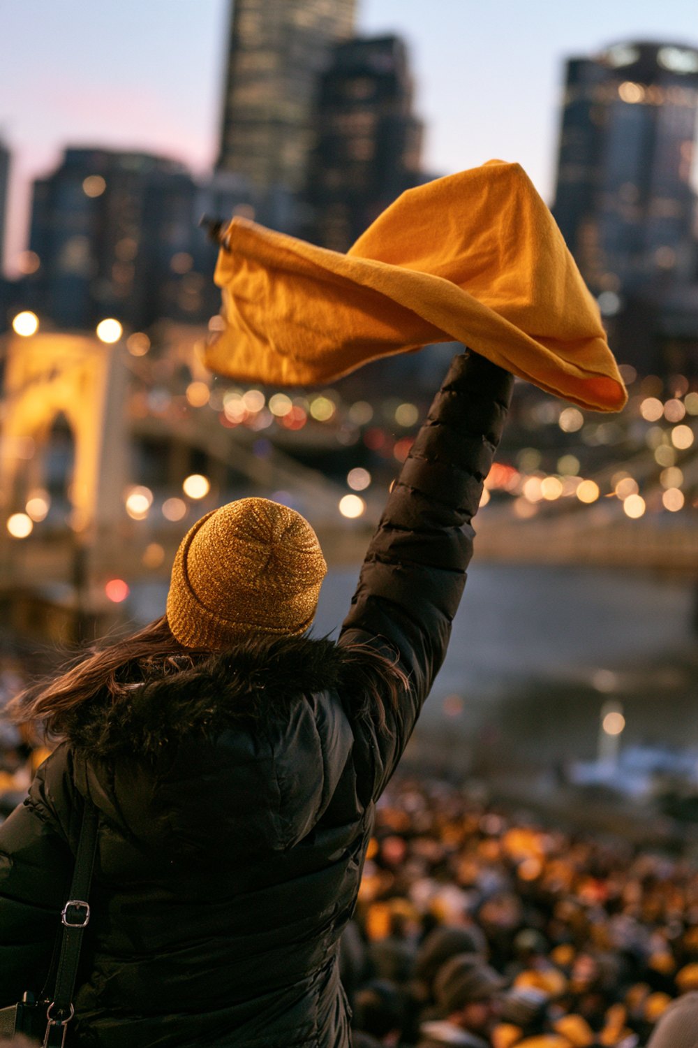 Fan from behind in a gold beanie and black coat waving a gold towel over the river at night