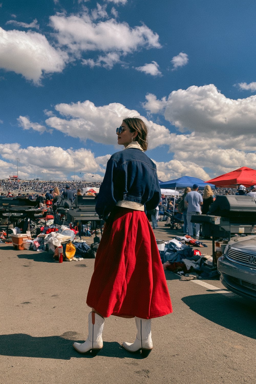 Woman seen from behind in a navy jacket, red midi skirt and white cowboy boots walking through a tailgate lot
