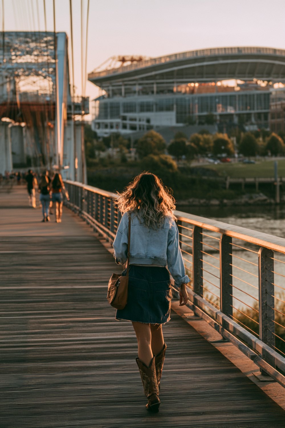 Woman seen from behind on a riverfront bridge walking toward a stadium in a denim jacket and boots
