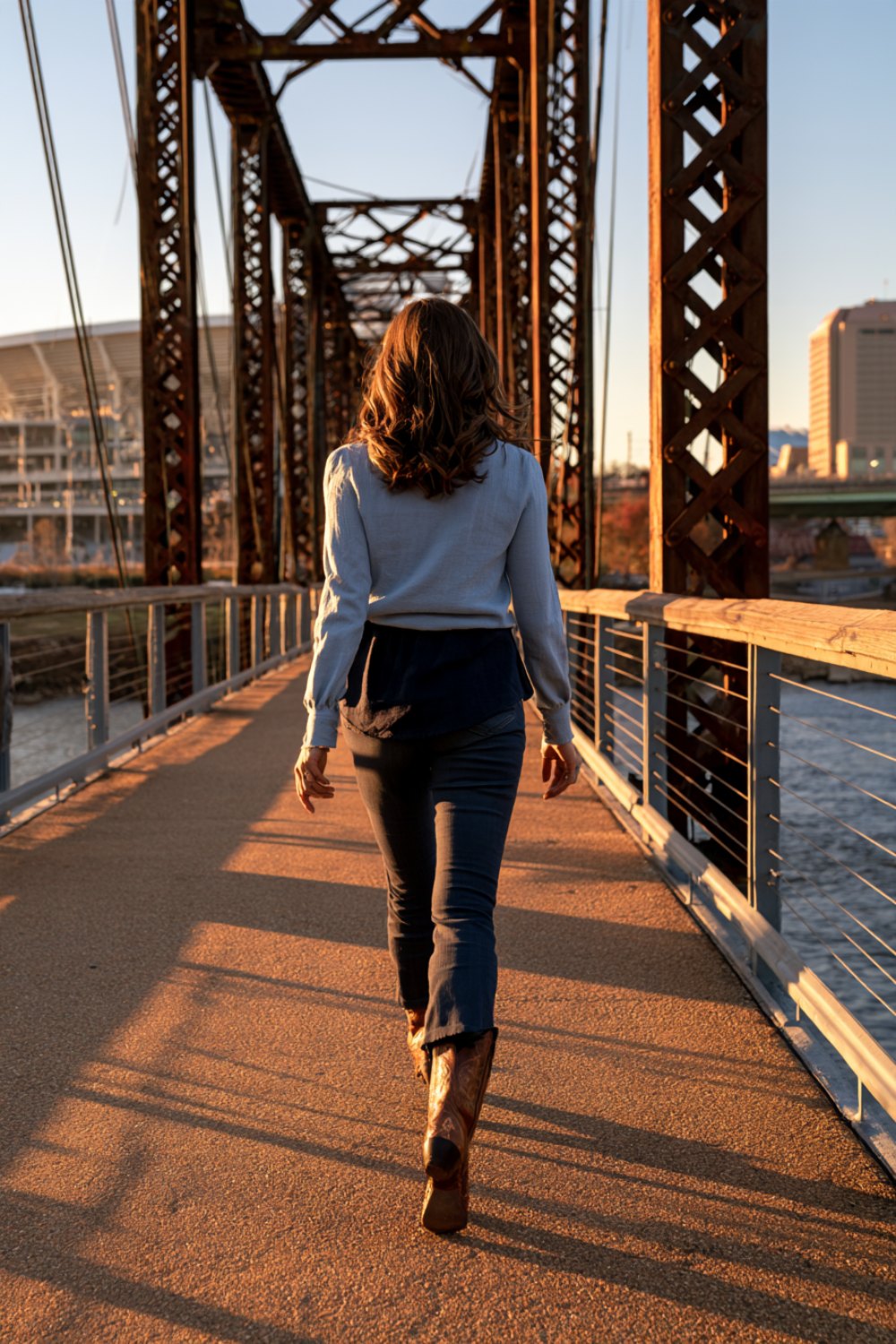 Woman from behind in a light blue top and boots walking across a steel truss pedestrian bridge at golden hour